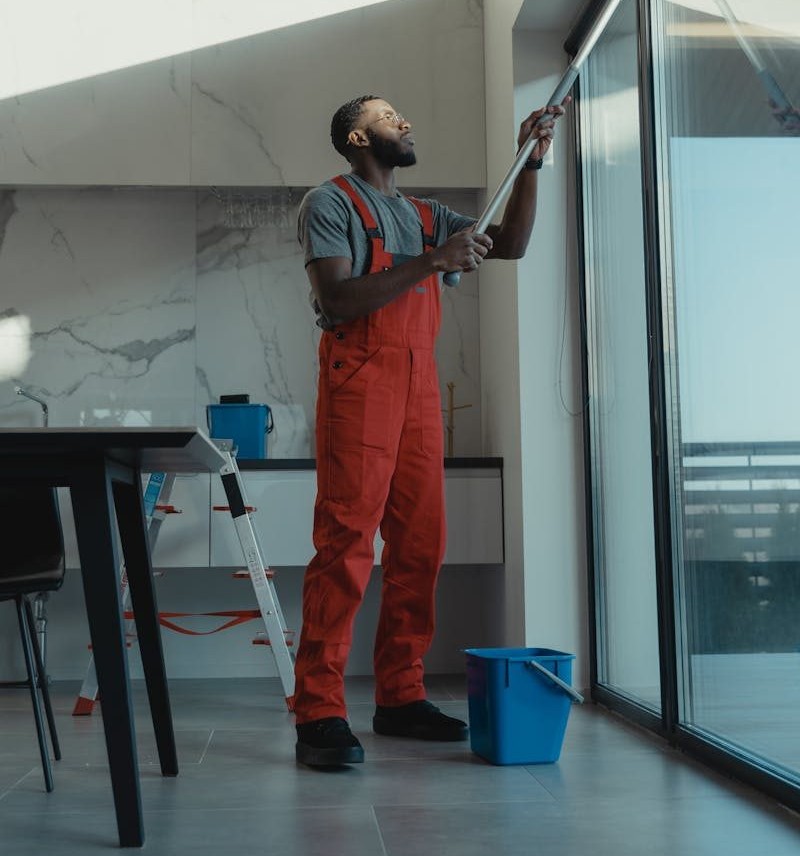 A man in coveralls cleans a large window in a stylish, modern home interior.